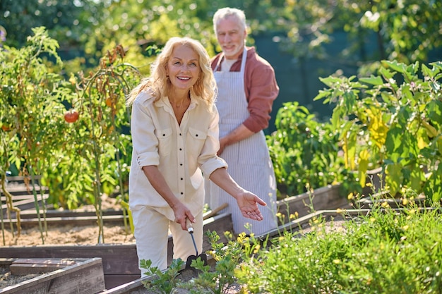 Hobby. joyful woman holding garden tools pointing to garden bed\
and man behind standing in grounds on sunny day