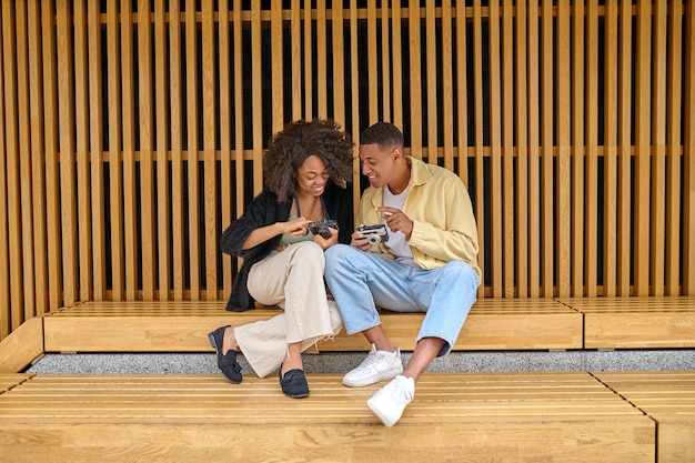 Hobby, photo. Dark-skinned young cheerful guy and girl sitting outdoors examining showing photos in camera communicating