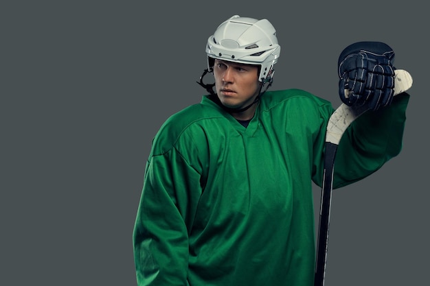 Hockey player wearing green protective gear and white helmet standing with the hockey stick on a gray background.