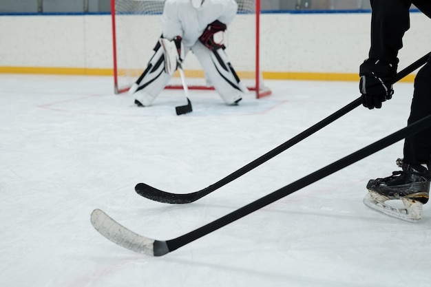 Hockeysticks vastgehouden door twee spelers in schaatsen, handschoenen en sportuniform op de achtergrond van de keeper die zich klaarmaakt om puck te vangen