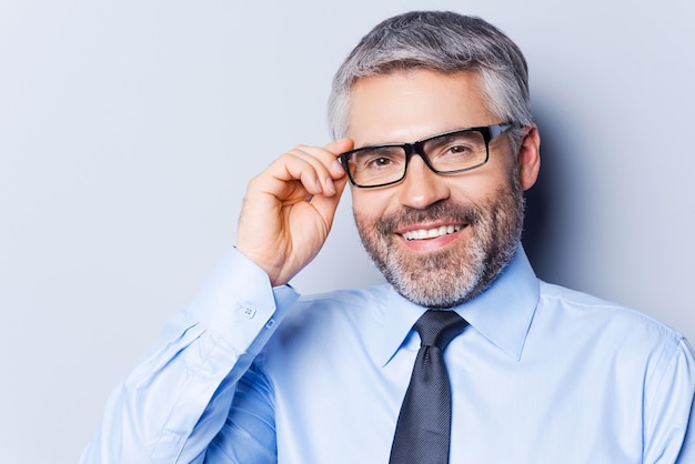 Hoe kan ik u helpen? Portret van een zelfverzekerde volwassen man in overhemd en stropdas die zijn bril aanpast en glimlacht terwijl hij tegen een grijze achtergrond staat