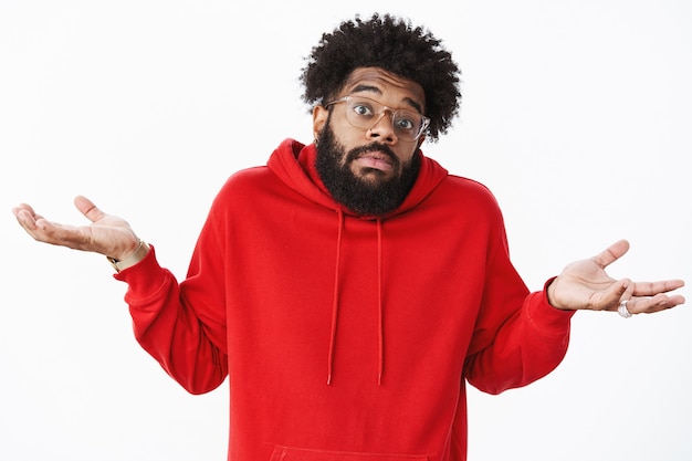 Hoe moet ik dat weten. Portret van een onwetende en onbewuste ongehinderde Afro-Amerikaanse mannelijke collega met baard in bril en rode hoodie, schouderophalend met handen zijwaarts gespreid, wenkbrauwen in verwarring optrekkend