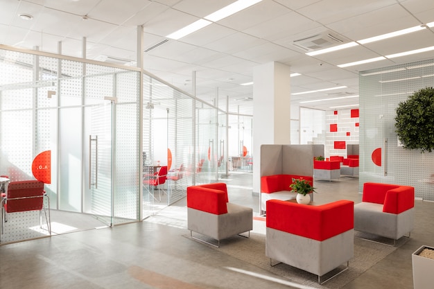 Hoek van modern kantoor met witte muren, grijze vloer, open plek met rode en witte fauteuils en kamers achter glazen wanden