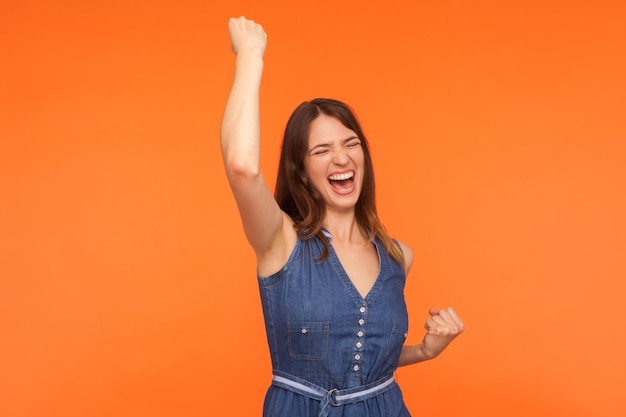 Hoera overwinning Gelukkig dolblij enthousiaste brunette vrouw in denim jurk dansen schreeuwen van opwinding bewonderen van succes gevoel als kampioen indoor studio opname geïsoleerd op oranje achtergrond