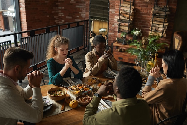 Hoge hoek portret van een multi-etnische groep van elegante jonge mensen bidden met gesloten ogen zittend aan tafel tijdens de viering van Thanksgiving,