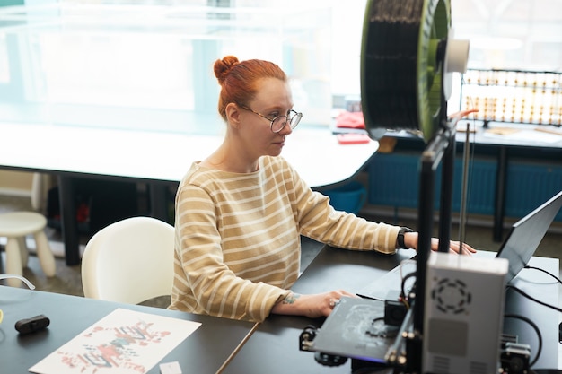 Hoge hoek portret van jonge roodharige vrouw met behulp van 3d-printer tijdens engineering klasse op de universiteit, kopieer ruimte