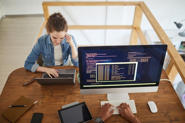 Hoge hoek portret van jonge vrouw met behulp van laptop tijdens het werken aan de balie in softwareontwikkelingsbureau met onherkenbare mannelijke collega code schrijven op computerscherm op voorgrond, kopie ruimte