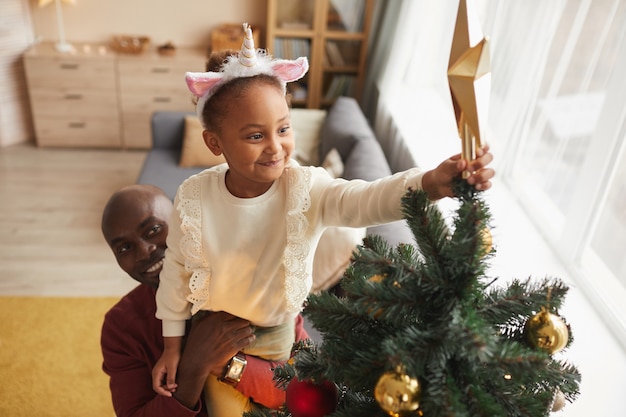 Hoge hoek portret van schattig Afrikaans-Amerikaans meisje ster zetten kerstboom met liefdevolle vader haar te helpen terwijl u geniet van vakantieseizoen thuis