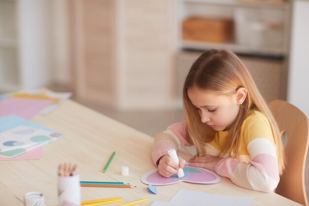 Hoge hoek portret van schattig klein meisje handgemaakte kaart maken voor moederdag zittend aan tafel in gezellig interieur, kopie ruimte