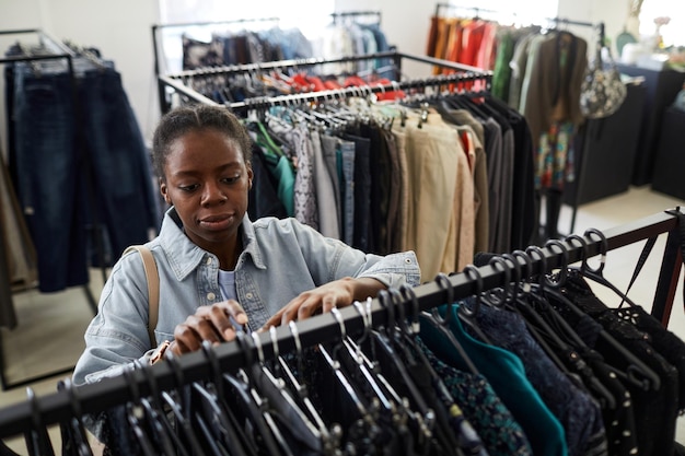 Hoge hoek portret van zwarte jonge vrouw die naar kleding kijkt tijdens het winkelen voor deals in kringloopwinkel