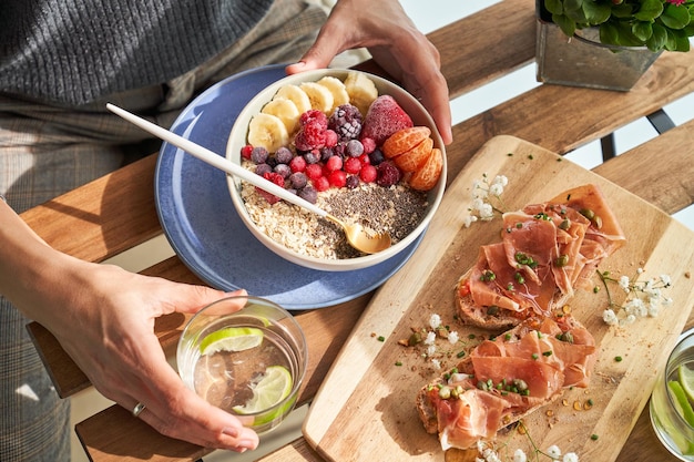 Hoge hoek van gewas, onherkenbare dame die limoenwater drinkt en heerlijk ontbijt eet met muesli en ham toast in zonnige ochtend op het terras van het restaurant