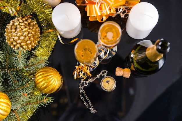 Hoge hoekmening van antieke zakhorloge op tafel omringd met fles champagne en glazen, witte kaarsen en groenblijvende takken versierd met gouden klatergoud Garland en kerstballen