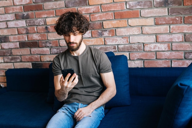 Hoge hoekmening van denkende, bebaarde jonge man die naar het scherm van de smartphone kijkt terwijl hij op draadloos internet surft op een apparaat dat thuis op de bank zit