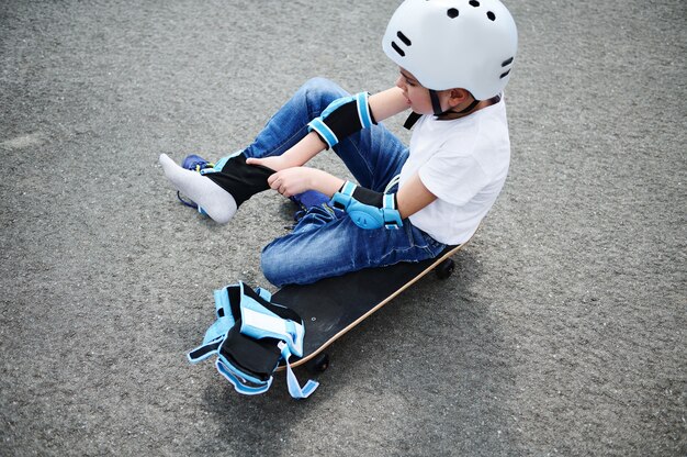 Hoge hoekmening van een sportieve jongen in veiligheidshelm zittend op een skateboard op speelplaatsasfalt en het aantrekken van beschermende kniebeschermers