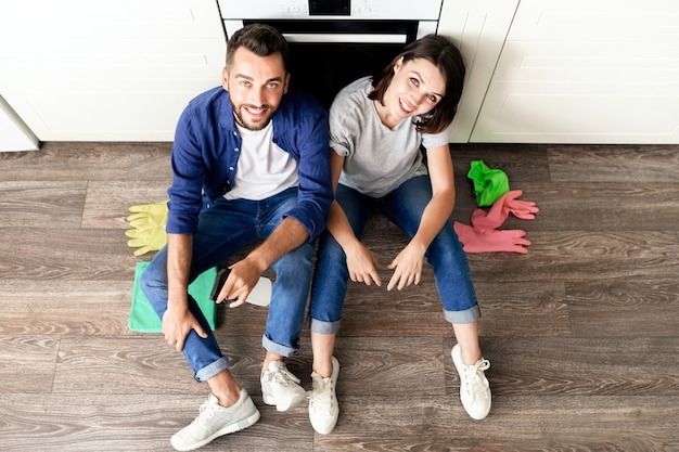 Hoge hoekmening van gelukkig zorgeloos jong koppel in casual kleding zittend op de vloer met rubberen handschoenen tijdens het rusten na het schoonmaken van huis