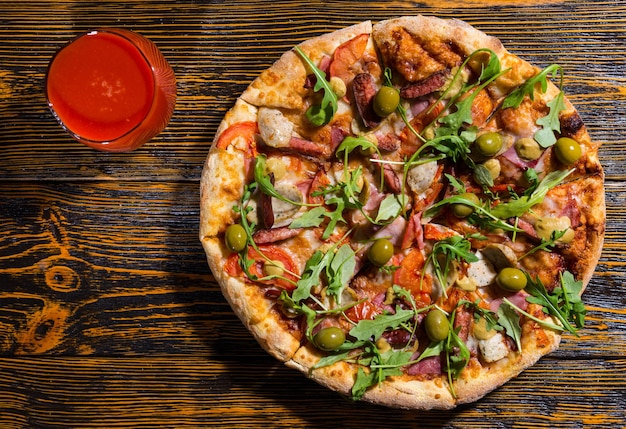 Hoge hoekmening van heerlijke pizza met verschillende toppings en kaas op houten tafel in de buurt van een glas met tomatensap