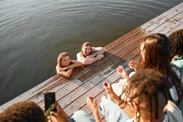 Hoge hoekmening van jong koppel zwemmen in het meer met een groep vrienden kijken vanaf de pier en filmin