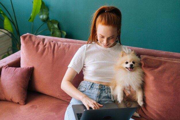 Hoge hoekmening van lachende jonge vrouw typen op laptopcomputer zittend op de bank knuffelen en strelen mooie miniatuur spitz huisdier hond. Gelukkige vrouwelijke freelancer die online op kantoor aan huis werkt.