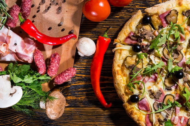 Hoge hoekmening van pizza met rucola en olijven op houten tafel, in de buurt van leugentomaten, champignons en snijplank met spek