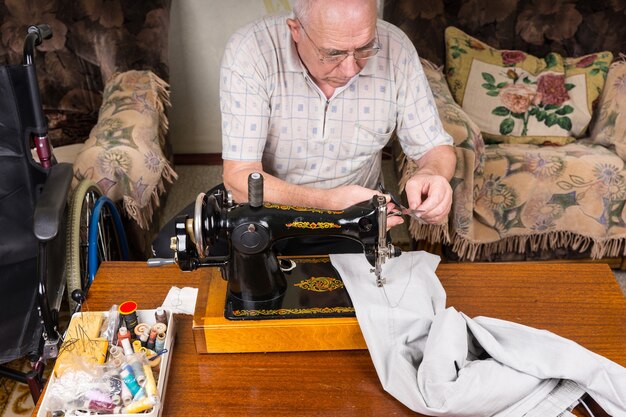 Hoge hoekmening van Senior Man herstellen broek met ouderwetse handmatige naaimachine thuis in woonkamer