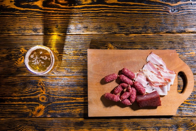 Hoge hoekmening van snijplank met worstjes, spek en vlees op houten tafel in de buurt van een glas bier