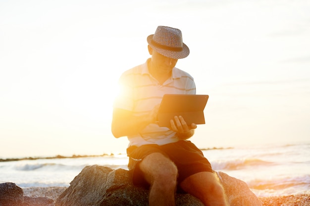 Hogere mens die aan zijn laptop op het strand tijdens zonsondergang werkt