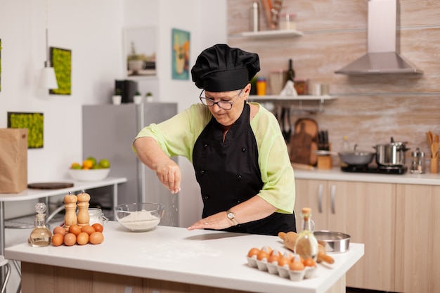 Hogere vrouw die bloem in huiskeuken voor bakkerijproducten verspreidt. Gelukkige bejaarde chef-kok met uniforme besprenkeling, zeven, zeven, zeven, zeven, rauwe ingrediënten, met de hand, bakken, zelfgemaakte, pizza.