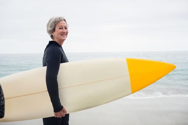 Hogere vrouw die in wetsuit een surfplank op het strand houdt