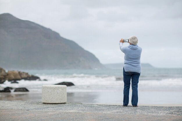 Hogere vrouw die landschap fotografeert die celtelefoon op het strand met behulp van