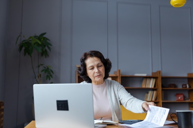 Hogere vrouw van middelbare leeftijd zit met laptop en papieren document peinzende oudere volwassen dame die papier leest
