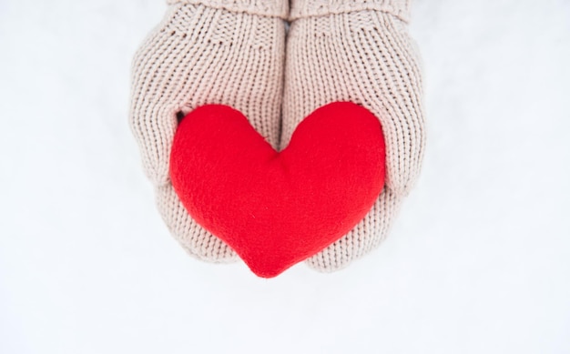 Hold red soft heart toy made of fabric in beige mittens against background of white snow