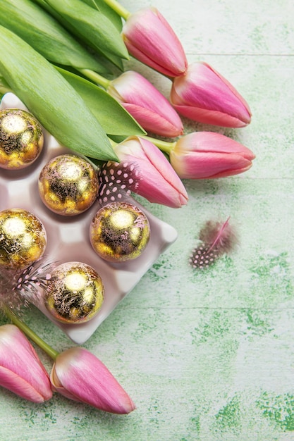 Holder with Easter eggs painted golden colors and pink tulips on a green wooden background