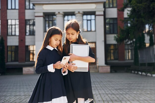 Две школьницы вместе держат книги на улице возле здания школы
