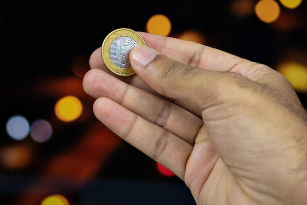 Holding a Brazilian money coin at a blurry lights background