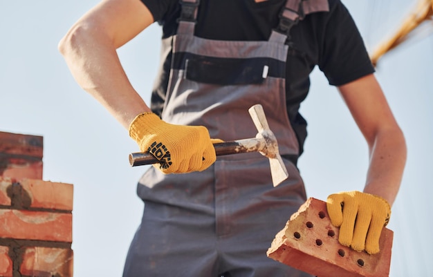 Holding brick and using hammer Construction worker in uniform and safety equipment have job on building