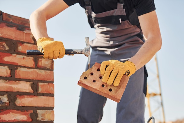 Holding brick and using hammer Construction worker in uniform and safety equipment have job on building