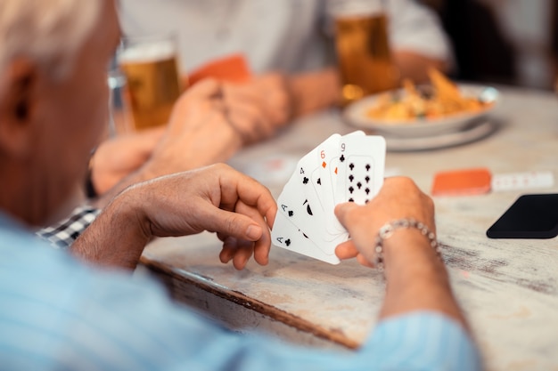 Holding cards. Close up of grey-haired retired man wearing bracelet holding cards