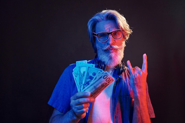 Holding cash Neon lighting Stylish modern senior man with gray hair and beard is indoors