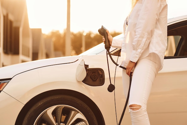 Photo holding charger young woman in white clothes is with her electric car at daytime