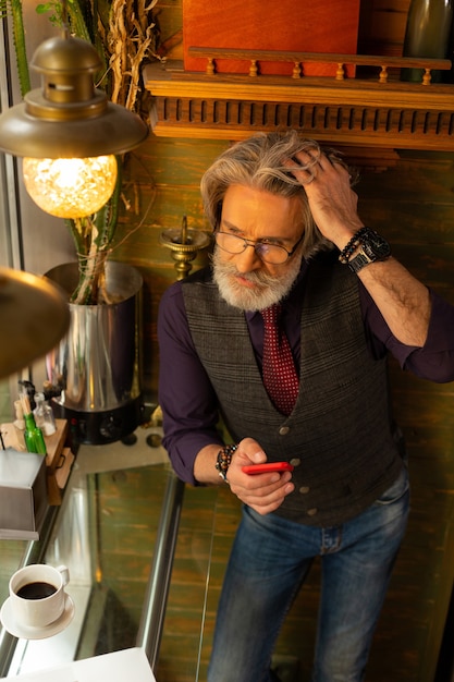 Photo holding correspondence. handsome adult man standing near the coffee shop table fixing his hair and thinking about his respond to a message.