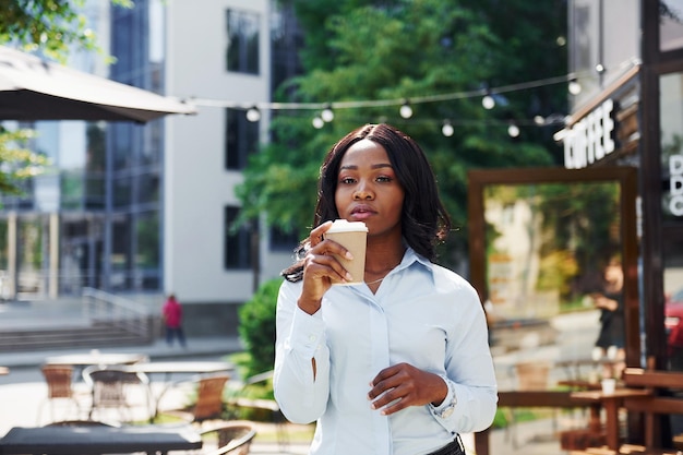 Holding cup of drink in hands Young afro american woman in white shirt outdoors in the city near green trees and against business building