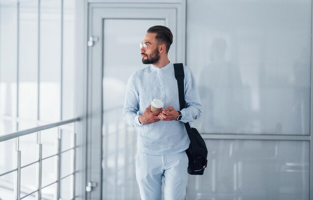 Holding cup with drink and black bag Young handsome man in formal clothes indoors in the office at daytime