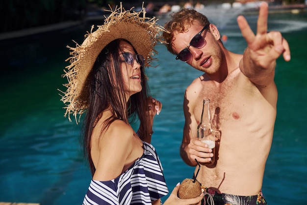 Holding drinks in hands Cheerful couple or friends together in swimming pool at vacation