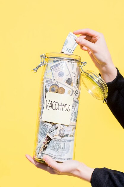 Photo holding a glass bottle full of money savings for the summer vacation on the yellow background