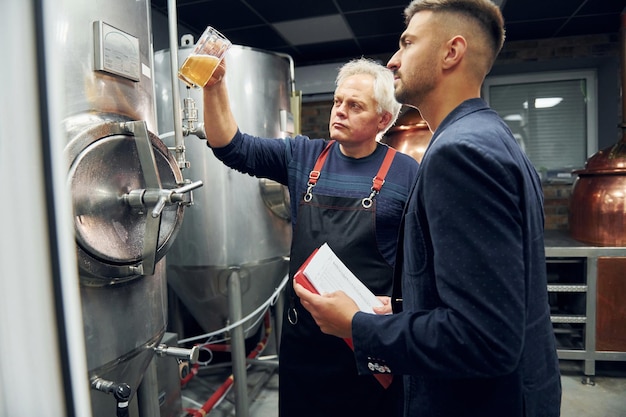 Holding glass senior man and young worker with document is\
standing in the storage beer room