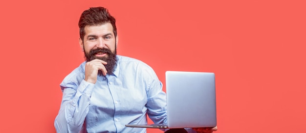 Holding laptop computer. Smiling handsome bearded man worker laptop. Happy young man sitting on the floor with and using laptop computer on red background. Young businessman using his laptop, pc.