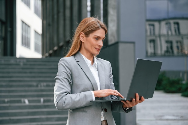 Holding laptop in hands standing Woman in formal clothes is outdoors near the business building