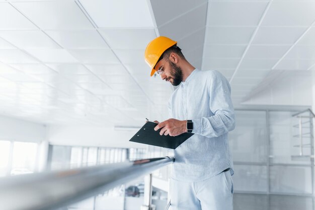 Holding notepad Engineer in white clothes and orange protective hard hat standing and working indoors