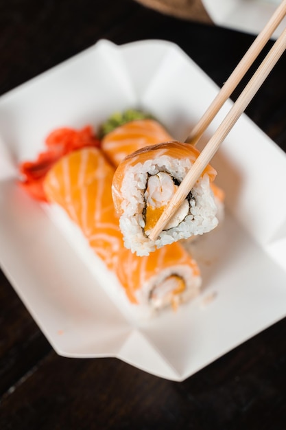 Holding philadelphia roll using chopstics for sushi. Roll with salmon, shrimp and mango. Japanese food delivery.