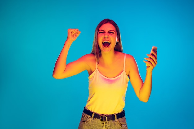 Holding phone, crazy happy winner, shocked. Caucasian woman's portrait on blue background in neon light. Beautiful model in casual. Concept of human emotions, facial expression, sales, ad. Copyspace.